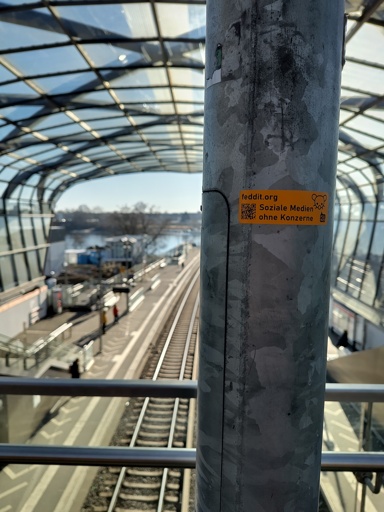 ein Feddit Sticker in kleinster-gemeinsamer-Nenner-Ausgang angeklebt an einer Säule mit einem Blick auf ein zwei gleisiges Gleisbett an einem hellen Tag ohne Wolken.